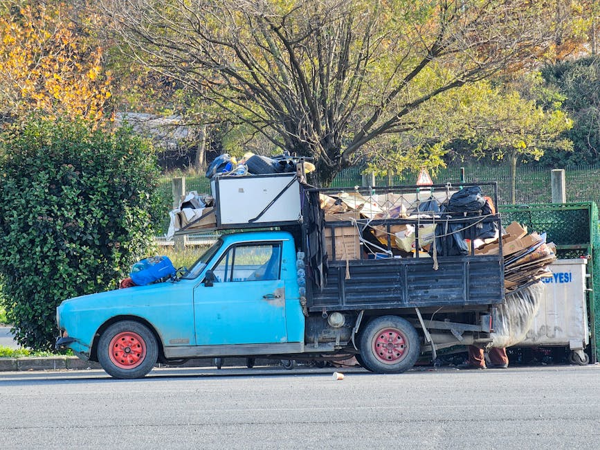 A small blue flatbed truck parked on the side of a street, loaded with a large amount of assorted rubbish and waste materials. The load includes cardboard boxes, black garbage bags, plastic containers, and miscellaneous debris stacked in the truck's open bed, secured with ropes. The truck has red-painted wheels and a slightly worn appearance, with the cabin's paint showing some signs of age. In the background, there is a large tree with green and autumn-colored leaves, along with a green metal fence and some foliage, indicating a suburban or residential area. The scene, captured in natural daylight, reflects an informal waste accumulation that may be associated with private rubbish collection or on-site clearance services, such as those provided by Waste Disposal Belgravia, involved in alternative waste handling in Belgravia, BELGRAVIA.