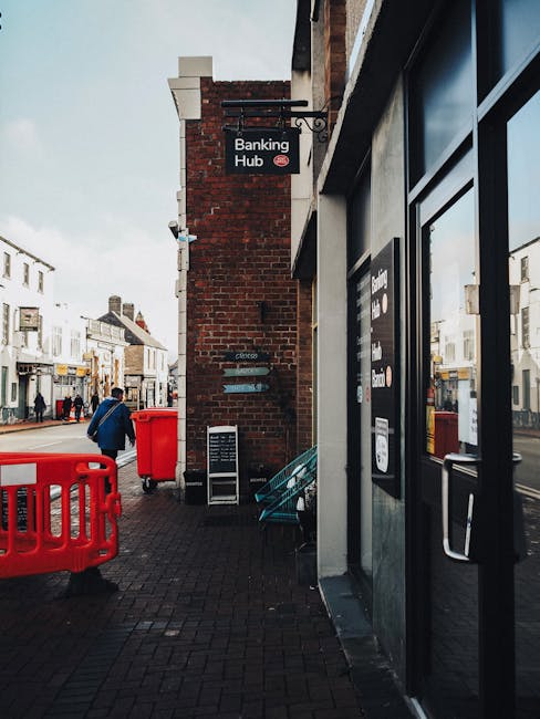 A street scene featuring a brick wall with a hanging sign labeled 'Banking Hub' attached to the side of a commercial building. The foreground shows a black door with glass panels adjacent to the brick wall, and nearby is a window with a black frame. There are red plastic barriers and a red waste bin positioned on the pavement, which is paved with dark bricks. In the background, pedestrians are walking along the street, and a row of shops with various storefronts and signs are visible, illuminated by natural daylight. The overall scene depicts a typical urban environment where private waste disposal services, like those provided by Waste Disposal Belgravia, might be involved in managing rubbish removal for local businesses. The image emphasizes the importance of on-site clearance and alternative waste handling solutions in busy commercial streets.