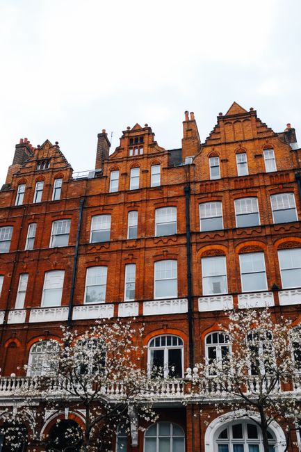 This image depicts a traditional red brick building with multiple floors, characteristic of Victorian architecture, located in an urban setting. The façade features rows of evenly spaced white-framed sash windows, some with decorative arches, and ornamental brickwork detailing above each window. The building's upper levels are topped with stepped gables and small chimneys, with a grey slate roof visible at the roofline. In the foreground, there are bare-branched trees with white blossom, suggesting early spring. The lower part of the building appears to have a small balcony or terrace area with a white balustrade. The overall scene presents a residential or mixed-use structure typical of Belgravia, with a clean, well-maintained appearance. The neutral lighting, possibly on an overcast day, highlights the warm tones of the brickwork, and the environment reflects a quiet, cityscape context, suitable for discussing alternative waste handling or private rubbish removal services by Waste Disposal Belgravia, especially relevant for maintaining such buildings' cleanliness and waste management needs.
