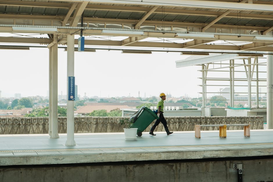 A worker wearing a yellow safety helmet and a green shirt is walking along an outdoor platform, pulling a large green wheeled rubbish bin behind him. The bin appears to be made of sturdy plastic with a textured surface, and it is slightly tilted as the individual moves it forward. There is a white plastic bucket placed on the platform near the bin, and several small, multicoloured plastic stools are arranged in a row towards the right side of the image. The platform is covered by a metal roof structure with visible beams and supports, providing shade and shelter. In the background, a cityscape with various buildings, rooftops, and trees is visible, indicating an urban environment. The scene is illuminated by natural daylight, highlighting the neutral tones of the platform, roof, and surrounding structures. This setting suggests a private waste collection activity, possibly part of an independent rubbish removal service, reflecting the context of alternative waste handling methods outside local authority collection, with Waste Disposal Belgravia operating in the background of such urban rubbish clearance scenarios.